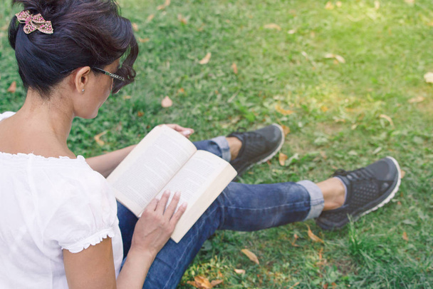 A girl is reading in the park. A concept of calm mood and relaxation in a park on the green grass. A middle-aged woman in blue jeans. - Foto, Imagem