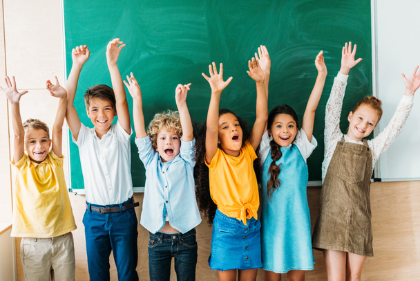 adorables escolares felices con las manos levantadas de pie delante de pizarra en blanco
 - Foto, Imagen