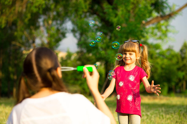 Young mother and little daughter playing in park with soap bubbles. Love family, parenthood, childhood - Foto, afbeelding