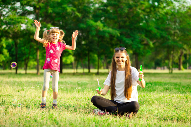 Young mother and little daughter playing in park with soap bubbles. Love family, parenthood, childhood - Valokuva, kuva