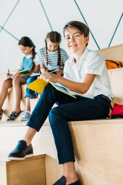 Not defterinde yazılı ve kamera arkasında oturan arkadaşlarından süre bakarak mutlu okul çocuğu - Fotoğraf, Görsel