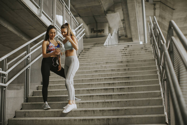 Kaksi naispuolista juoksijaa, jotka etsivät älypuhelimella lepoa lenkkeilyn jälkeen kaupunkiympäristössä
 - Valokuva, kuva