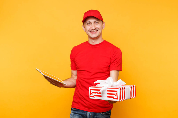 Delivery man in red uniform isolated on yellow orange background. Male employee courier in cap t-shirt holding red present box with ribbon, gift, using tablet pc computer. Service concept. Copy space - Foto, afbeelding