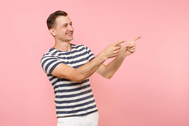 Ritratto di giovane uomo sorridente che indossa una t-shirt a righe che punta le dita da parte su uno spazio di copia isolato su uno sfondo rosa pastello di tendenza. Persone emozioni sincere concetto di stile di vita. Area pubblicitaria
 - Foto, immagini