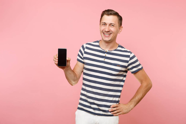 Portrait of man in striped t-shirt showing mobile phone camera with blank black empty screen copy space isolated on trending pastel pink background. People sincere emotions concept. Advertising area - Photo, Image