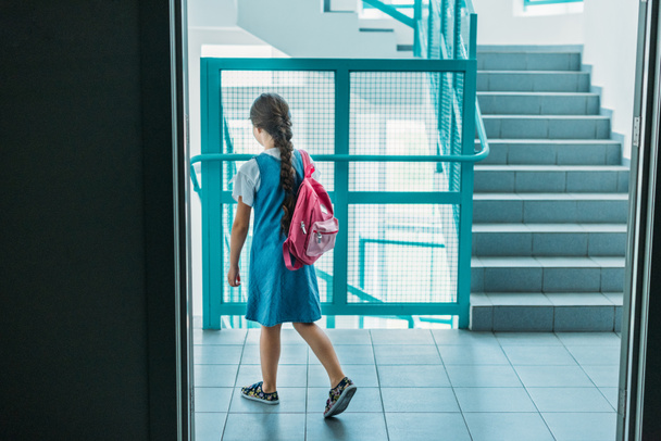 Rückansicht einer kleinen Schülerin in Kleid, die mit Rucksack auf dem Schulflur spaziert - Foto, Bild