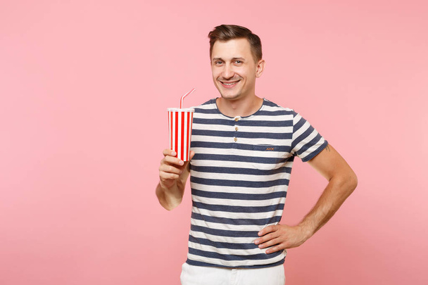 Portrait of young man wearing striped t-shirt holding plastic cup of soda, red glass cola isolated on trending pastel pink background. People youth sincere emotions lifestyle concept. - Fotoğraf, Görsel