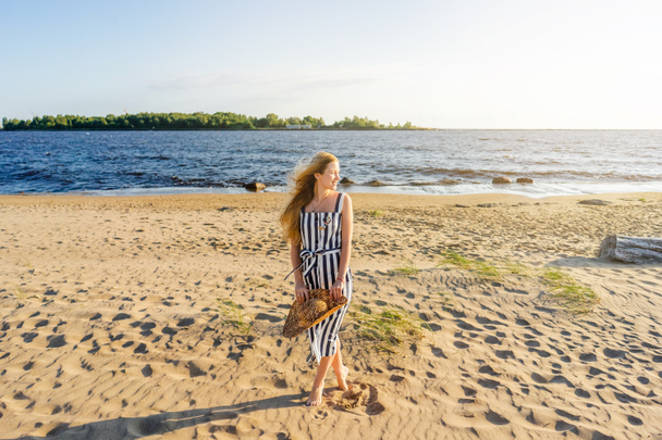 schöne lächelnde Frau mit Strohhut, die allein am Sandstrand mit Meer im Hintergrund steht - Foto, Bild