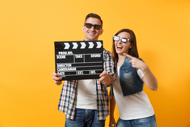 Giovane coppia sorridente uomo donna in occhiali 3d guardare film in data indicando dito indice sul classico film nero rendendo clapperboard isolato su sfondo giallo. Emozioni nel concetto di cinema
 - Foto, immagini