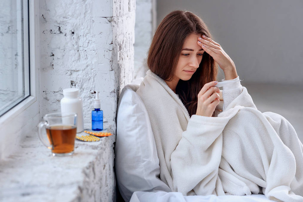 Health care concept. Beautiful ill woman with headache, sore throat and fever covered in blanket feeling sick, measuring body temperature with thermometer and holding her head. Sickness and illness. - Foto, Imagen