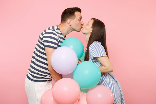Portrait of young happy kissing couple in love. Woman and man in blue clothes celebrating birthday holiday party on pastel pink background with colorful air balloons. People sincere emotions concept - 写真・画像