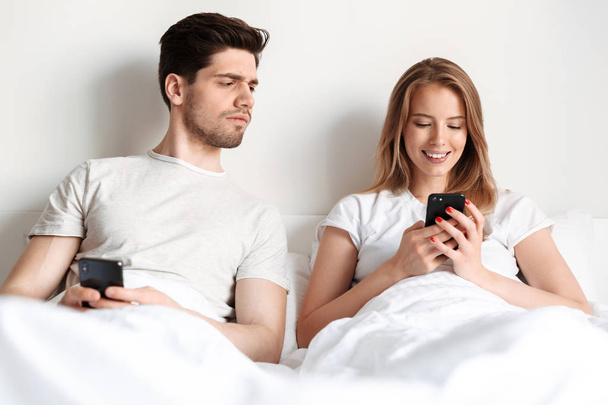 Image of happy young woman lies in bed using smartphone chatting while her husband looking to her phone. - Foto, immagini