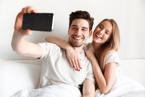 Изображение молодой веселой любящей пары лежит в постели сделать селфи на смартфоне глядя в сторону
. - Фото, изображение