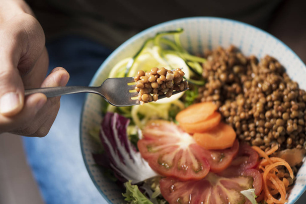 high angle view of a young caucasian man, wearing blue jeans, eating a buddha bowl made with lentils, carrot, zucchini, lettuce and tomato - Photo, Image