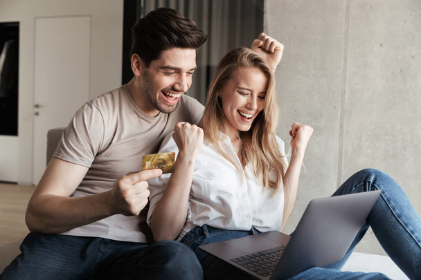 Photo of young happy loving couple in home indoors on sofa using laptop computer holding credit card showing winner gesture. - Foto, Imagen