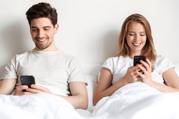 Image of happy young loving couple lies in bed using mobile phones chatting. - Foto, afbeelding