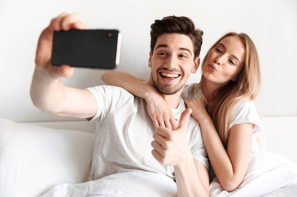 Image of young cheerful loving couple lies in bed take a selfie by smartphone looking aside showing thumbs up gesture. - Foto, imagen