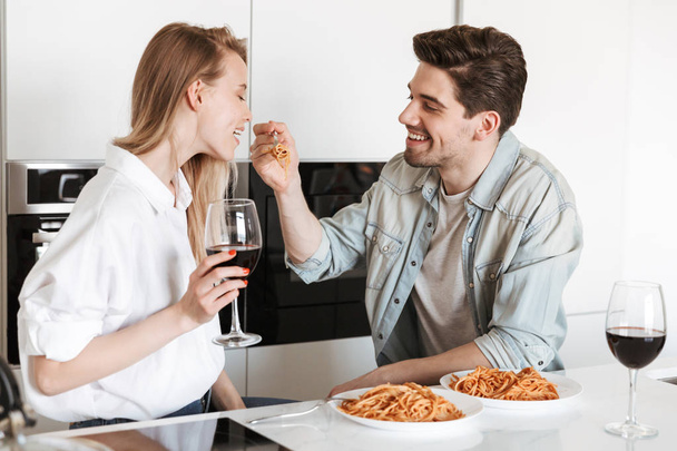 Image of young happy loving couple in kitchen have a dinner drinking wine eating spaghetti. - Фото, зображення