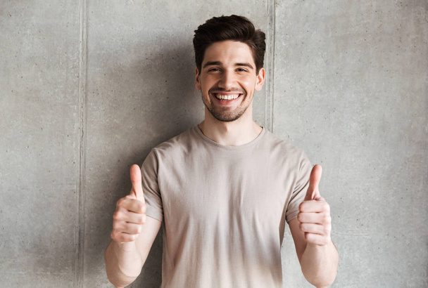 Image of happy young man standing over grey wall background looking camera showing thumbs up. - Foto, imagen