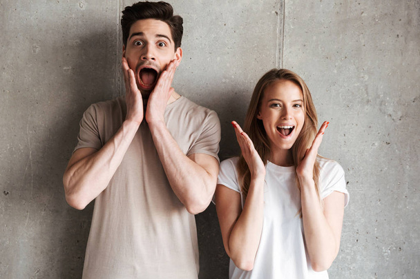 Photo of young excited people man and woman in basic clothing screaming and raising hands at face isolated over concrete gray wall indoor - Zdjęcie, obraz
