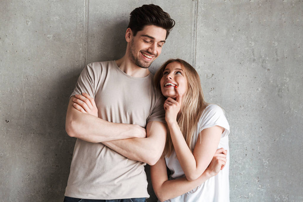 Portrait of content young people man and woman in basic clothing smiling and looking at each other isolated over concrete gray wall indoor - Photo, Image