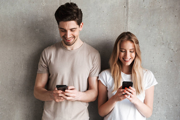 Photo of positive people man and woman smiling while both using mobile phones isolated over concrete gray wall indoor - Fotó, kép