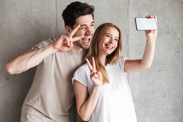 Image of beautiful couple man and woman taking selfie photo on smartphone while showing peace sign isolated over concrete gray wall indoor - Foto, imagen