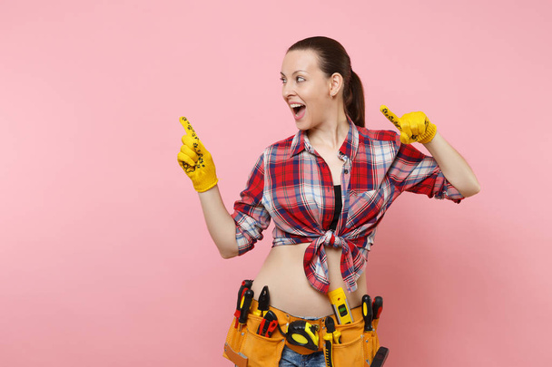 Strong young fun handyman woman in plaid shirt, denim shorts, yellow gloves, kit tools belt full of variety useful instruments isolated on pink background. Female doing male work. Renovation concept - Foto, imagen