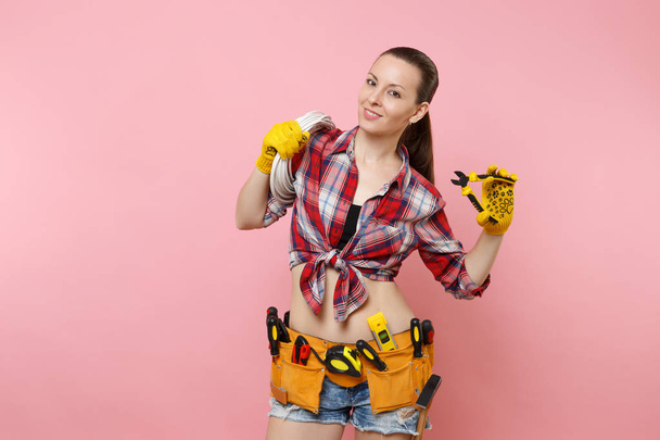Young excited handyman woman in plaid shirt, denim shorts, kit tools belt full of variety instruments holding electric power cabel isolated on pink background. Female in male work. Renovation concept - Photo, Image