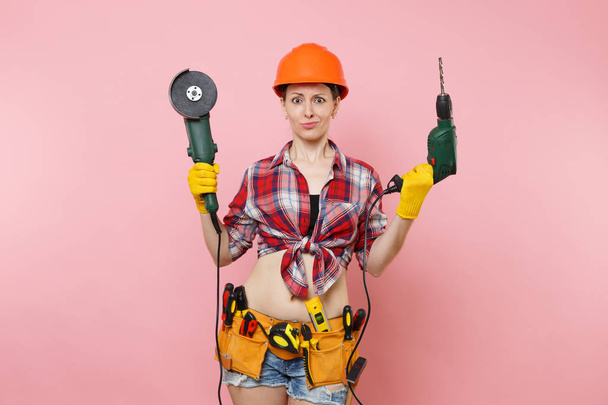 Energy fun handyman woman in gloves, protective orange helmet, kit tools belt full of instruments holding power saw electric drill isolated on pink background. Female in male work. Renovation concept - Photo, image