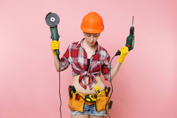 Energy fun handyman woman in gloves, protective orange helmet, kit tools belt full of instruments holding power saw electric drill isolated on pink background. Female in male work. Renovation concept - Photo, Image
