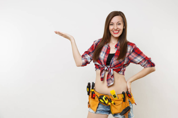 Strong young handyman woman in plaid top shirt, denim shorts, kit tools belt full of variety useful instruments isolated on white background. Female doing male work. Renovation and occupation concept - Photo, Image