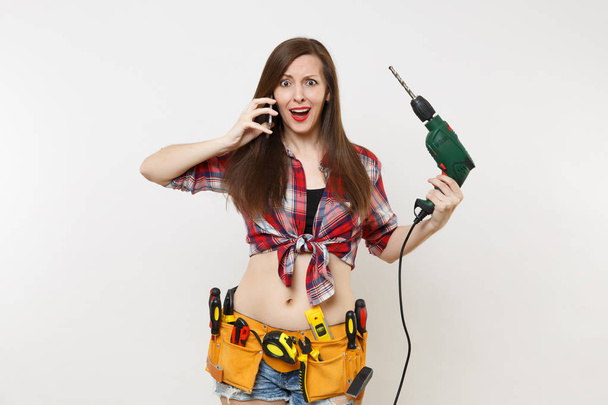 Young energy handyman woman in plaid shirt, kit tools belt full of instruments talking on mobile phone, hold power electric drill isolated on white background. Female in male work. Renovation concept - Foto, afbeelding