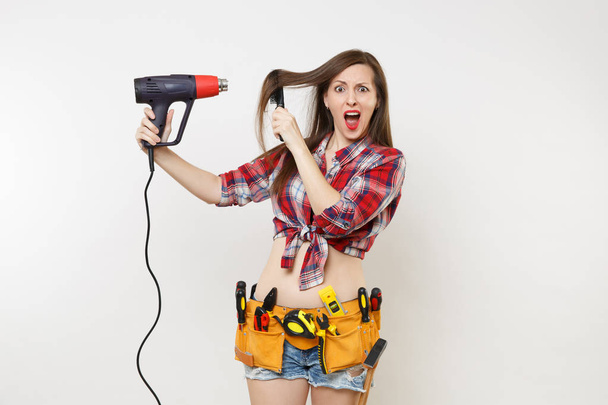 Silly mad strange fun handyman woman in plaid shirt, denim shorts, kit tools belt full of instruments dry hair with power heat gun isolated on white background. Female male work. Renovation concept - Foto, Imagem