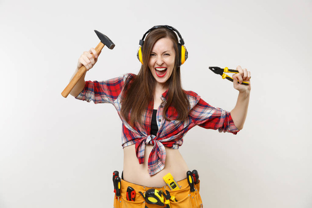 Strong fun young handyman woman in plaid top shirt, denim shorts, noise insulated headphones, kit tools belt full of instruments isolated on white background. Female in male work. Repair home concept - Photo, image
