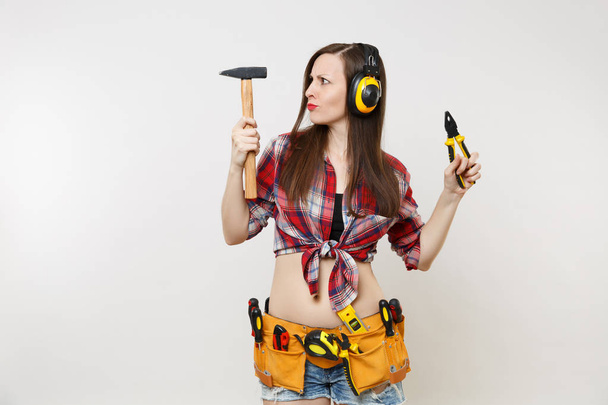 Strong fun young handyman woman in plaid top shirt, denim shorts, noise insulated headphones, kit tools belt full of instruments isolated on white background. Female in male work. Repair home concept - Valokuva, kuva