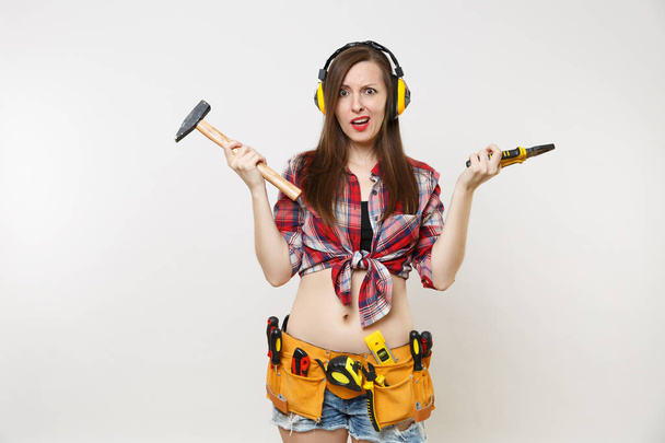 Angry emotional woman in tartan shirt, denim shorts, noise insulated headphones, kit tools belt full of instruments isolated on white background. Female in male work. Renovation occupation concept - Photo, Image