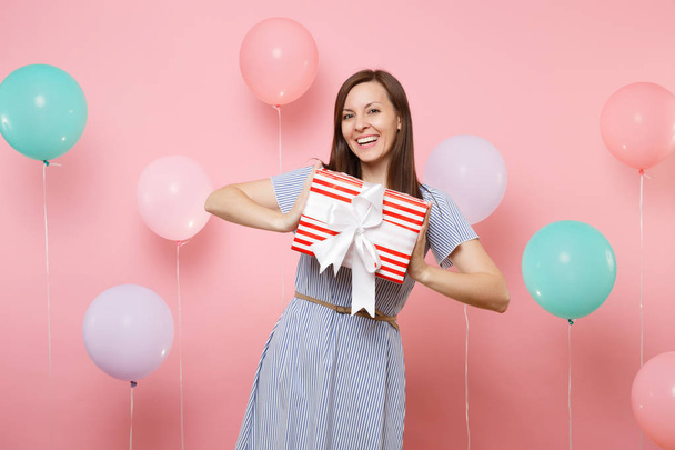 Portrait of beautiful smiling young woman in blue dress holding red box with gift present on pastel pink background with colorful air balloons. Birthday holiday party, people sincere emotions concept - 写真・画像