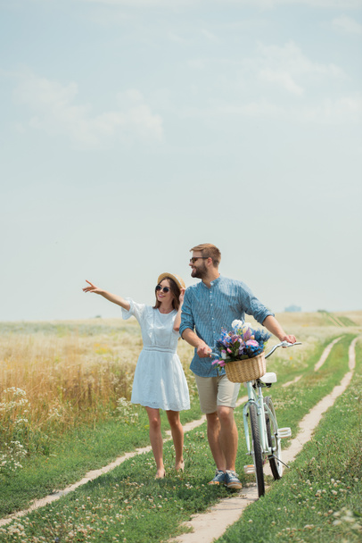 smiling couple in sunglasses with retro bicycle in summer field with wild flowers - Photo, Image