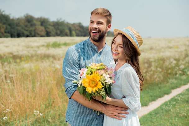 portrait of smiling man hugging girlfriend with bouquet of wild flowers in summer field - Fotó, kép