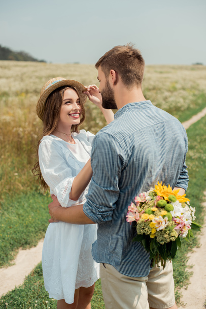 hombre escondiendo ramo de flores silvestres para la novia sonriente detrás de vuelta en el campo de verano
 - Foto, Imagen