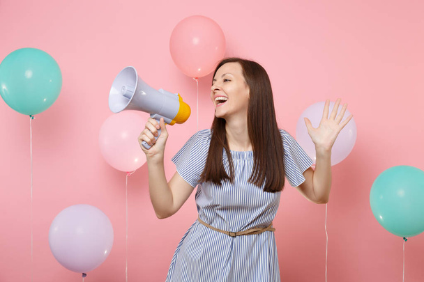Portrait of attractive young joyful woman in blue dress holding megaphone spreading hands on pastel pink background with colorful air baloons. Birthday holiday party, people sincere emotions concept - Photo, Image