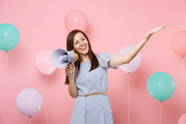 Портрет улыбающейся молодой красивой женщины в синем платье, держащей мегафон, указывающий на руку на розовом фоне с красочными воздушными шарами. День рождения праздник, люди искренние эмоции концепции
 - Фото, изображение