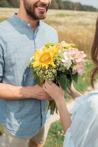 cropped shot of cheerful man presenting bouquet of wild flowers to girlfriend in summer field - Fotografie, Obrázek