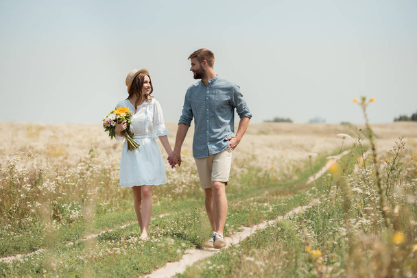 smiling woman in white dress with bouquet of wild flowers walking together with boyfriend in field - Photo, image