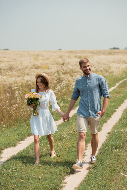 woman in white dress with bouquet of wild flowers walking together with boyfriend in field - Foto, Imagem