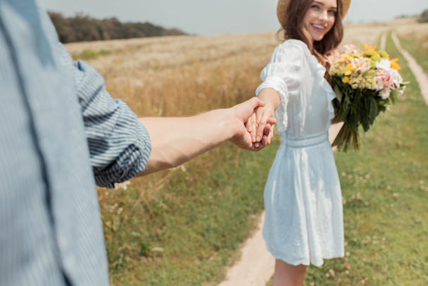 vista parcial de la mujer sonriente con ramo de flores silvestres y novio de la mano en el campo
 - Foto, Imagen