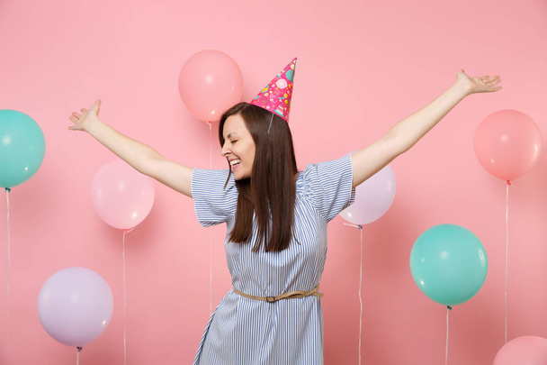 Porträt einer lachenden jungen Frau mit geschlossenen Augen in Geburtstagsmütze und blauem Kleid, die die Hände auf rosa Hintergrund mit bunten Luftballons ausbreitet. Geburtstagsparty, Menschen aufrichtige Emotionen Konzept - Foto, Bild