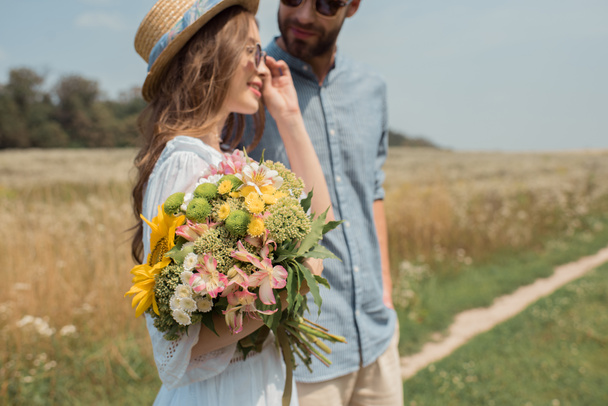 vista lateral de mujer sonriente con ramo de flores silvestres y novio cerca en el campo
 - Foto, Imagen