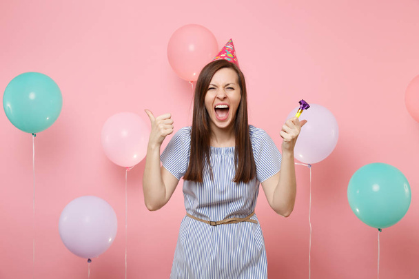 Portrait of crazy young woman with party whistle in birthday hat and blue dress spreading hands screaming on pink background with colorful air baloons. Birthday holiday party, people sincere emotions - Foto, Bild